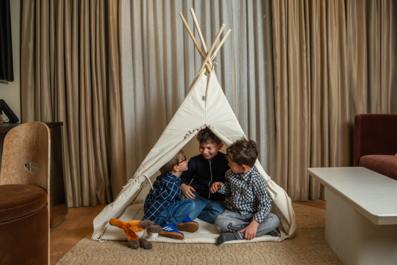 Three young boys laughing together inside a white play teepee set up in a modern luxury hotel suite.