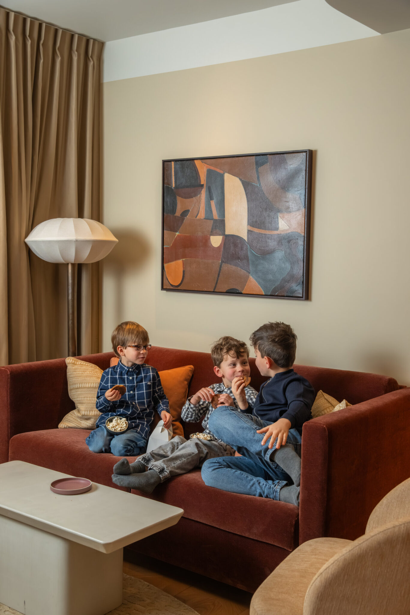 Three boys snacking and laughing together on a rust-red velvet sofa in a stylish luxury hotel suite.