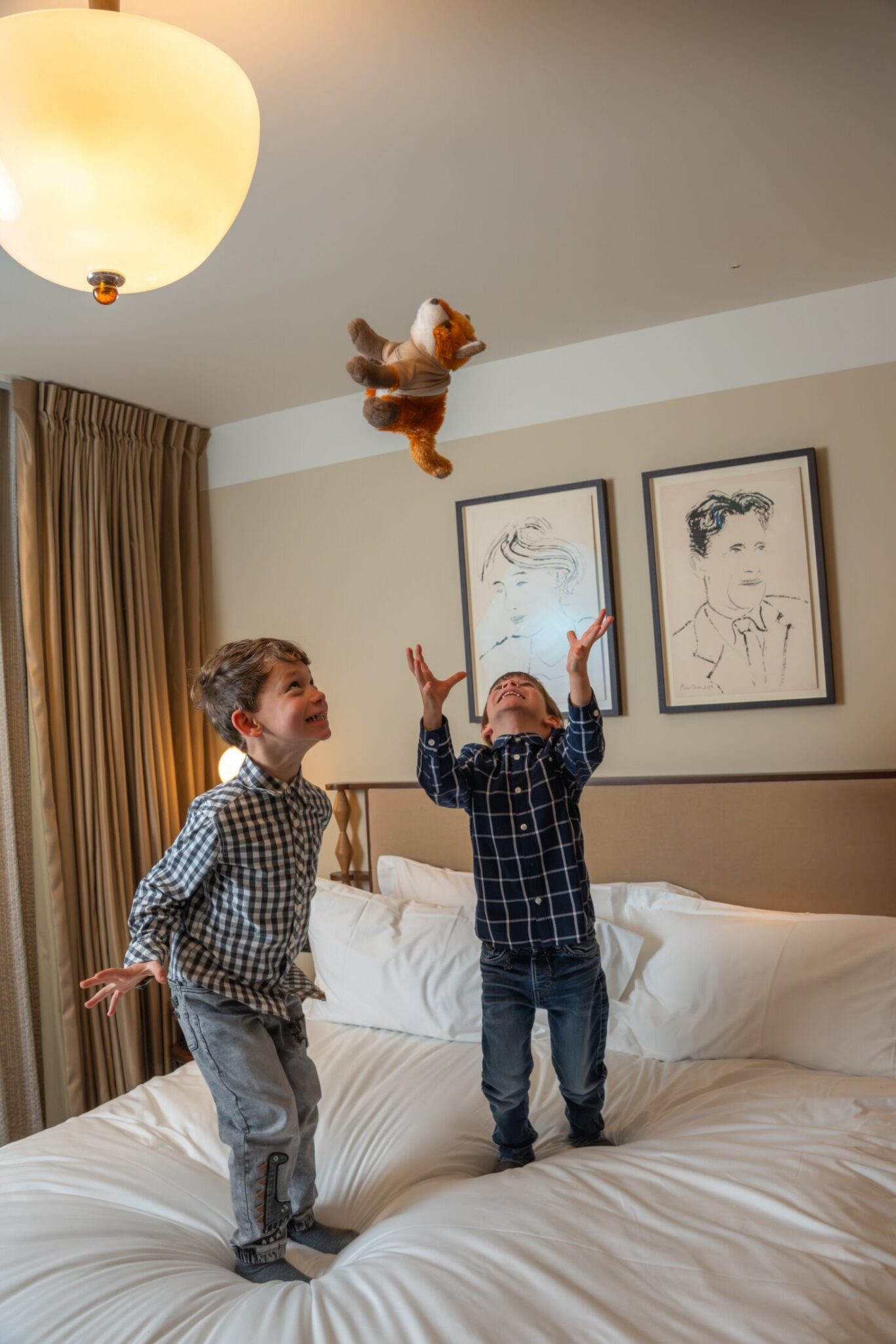 Two smiling boys standing on a hotel bed tossing a stuffed animal toy in the air in a warmly lit luxury room.