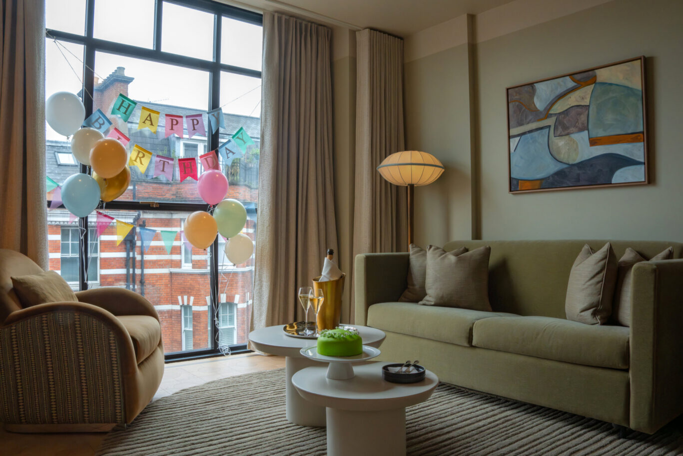 Luxury hotel suite living room decorated with birthday balloons, bunting, champagne, and green cake on white coffee tables.