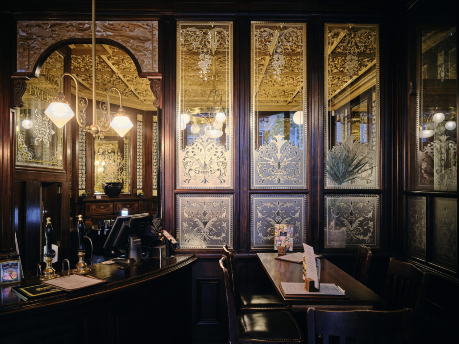 Ornate Victorian pub interior with etched glass screens, gilded panels, dark mahogany woodwork, and brass globe pendant lights.