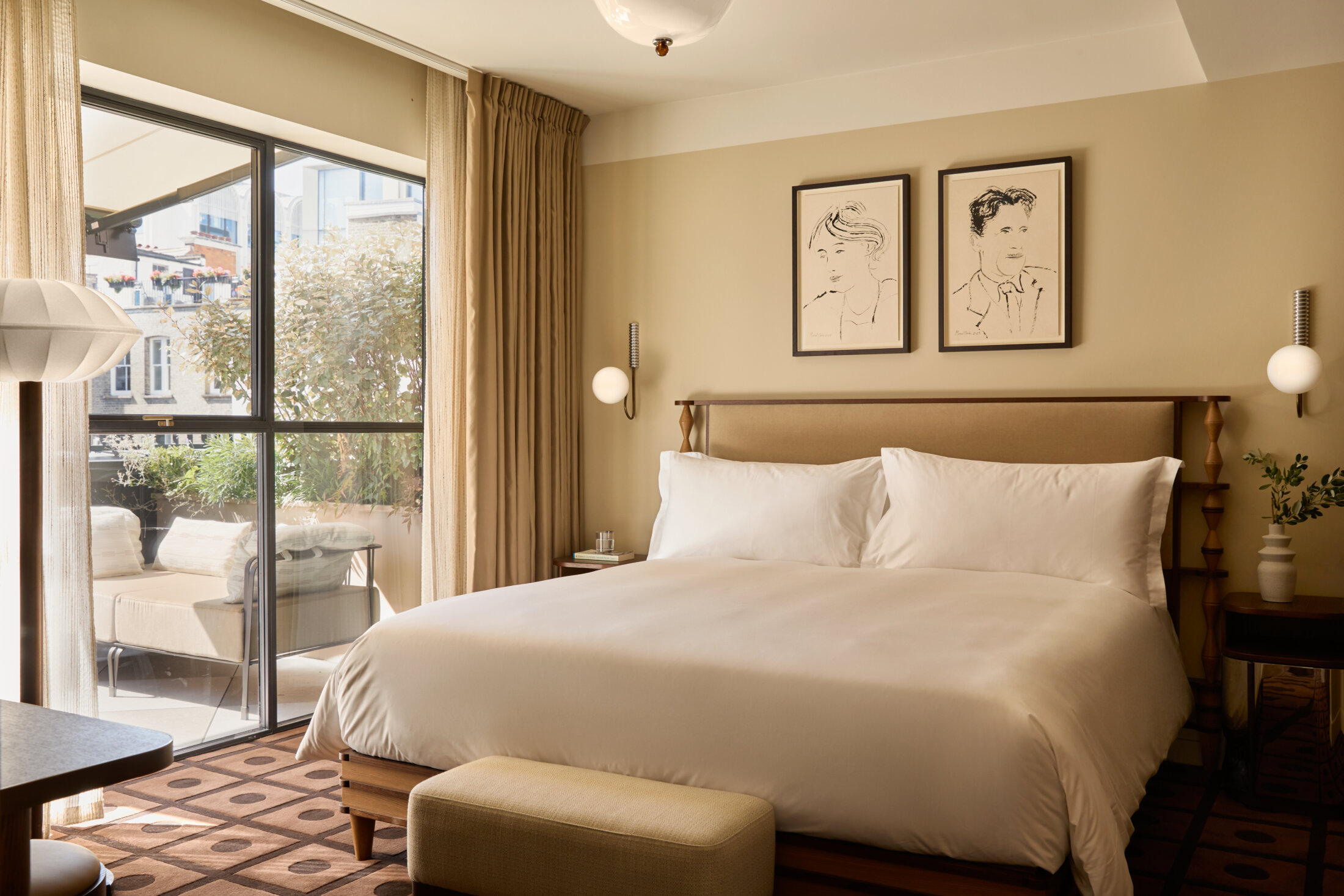 Sunlit luxury hotel bedroom with white king bed, terrace access, warm beige walls, and framed portrait sketches.